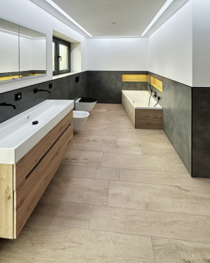 Modernes Badezimmer mit anthrazitfarbenen Wandfliesen, Holzfliesenboden, Waschtischunterschrank aus Holz, großem Doppelwaschtisch, schwarz-matte Armaturen, Toilette, Bidet, Badewanne und barrierefreier Dusche mit Regenduschsystem für stilvolles und funktionales Design.