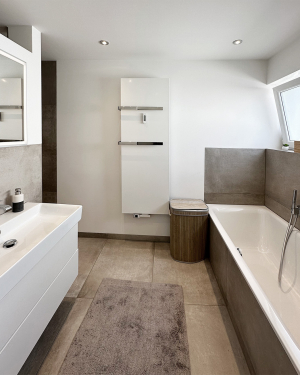 Badezimmer mit betonfarbenen Fliesen, Badewanne, WC, bodentiefer Dusche mit Regenduschsystem, Waschbecken mit weißem Waschtischunterschrank, Chrom-Armaturen und einem eleganten Wandheizkörper für zeitloses, funktionales Design und stilvolles Wohlfühlbad. Petri Heizung &amp; Sanitär, Mühlheim am Main