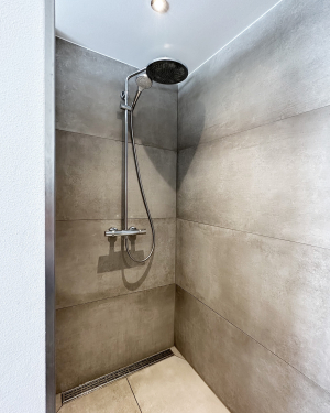 Badezimmer mit betonfarbenen Fliesen, Badewanne, WC, bodentiefer Dusche mit Regenduschsystem, Waschbecken mit weißem Waschtischunterschrank, Chrom-Armaturen und einem eleganten Wandheizkörper für zeitloses, funktionales Design und stilvolles Wohlfühlbad. Petri Heizung &amp; Sanitär, Mühlheim am Main
