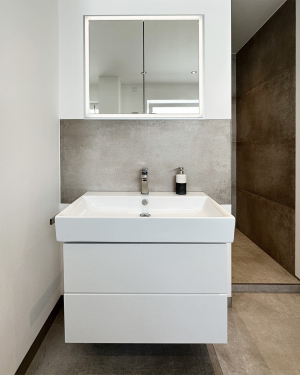 Badezimmer mit betonfarbenen Fliesen, Badewanne, WC, bodentiefer Dusche mit Regenduschsystem, Waschbecken mit weißem Waschtischunterschrank, Chrom-Armaturen und einem eleganten Wandheizkörper für zeitloses, funktionales Design und stilvolles Wohlfühlbad. Petri Heizung &amp; Sanitär, Mühlheim am Main